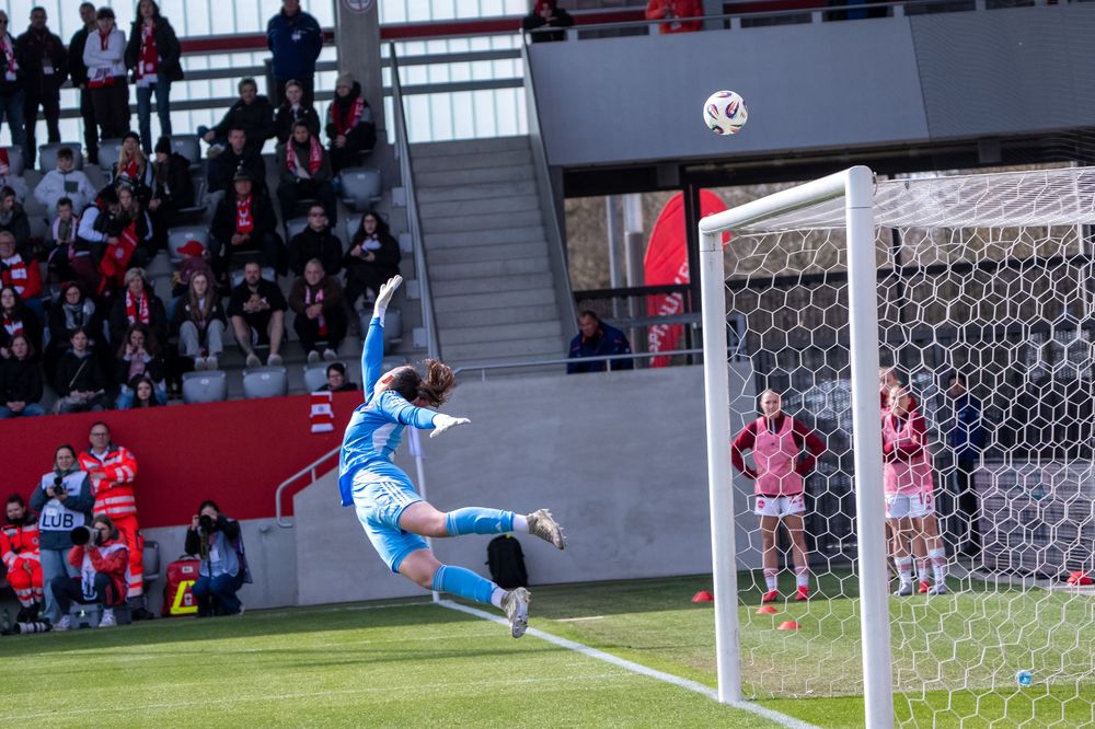 Soccer goalkeeper diving to make a save during a match, with spectators watching from the stands behind the goal.