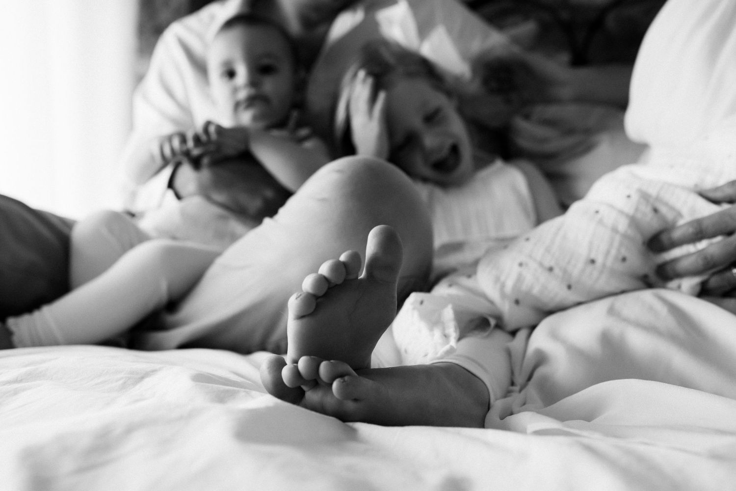 Black and white family photo of parents and children laughing together on a bed, baby feet in foreground.