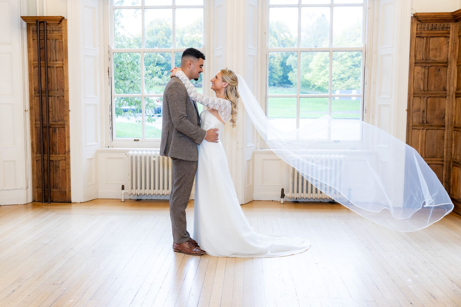 Bride and groom share a romantic embrace in an elegant white ballroom, with her veil flowing gracefully behind her.