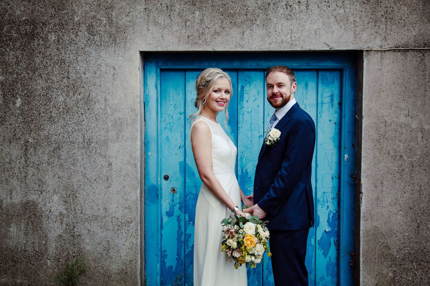 Bride and groom posing together in front of a rustic blue door on their wedding day.