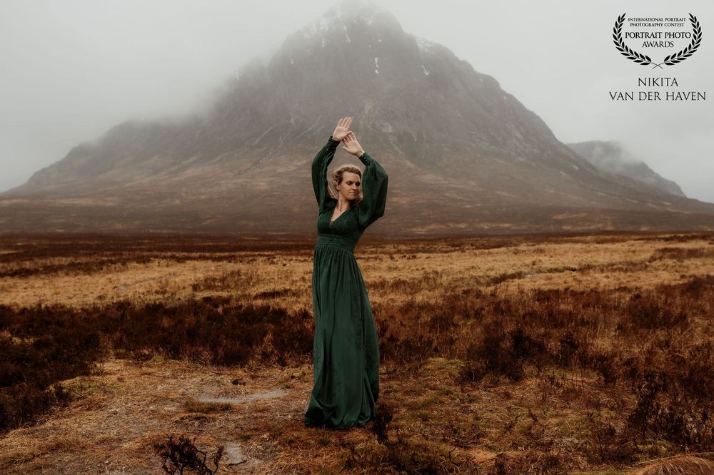 Woman in green dress dancing before misty mountain landscape in Scottish Highlands, award-winning portrait photography.