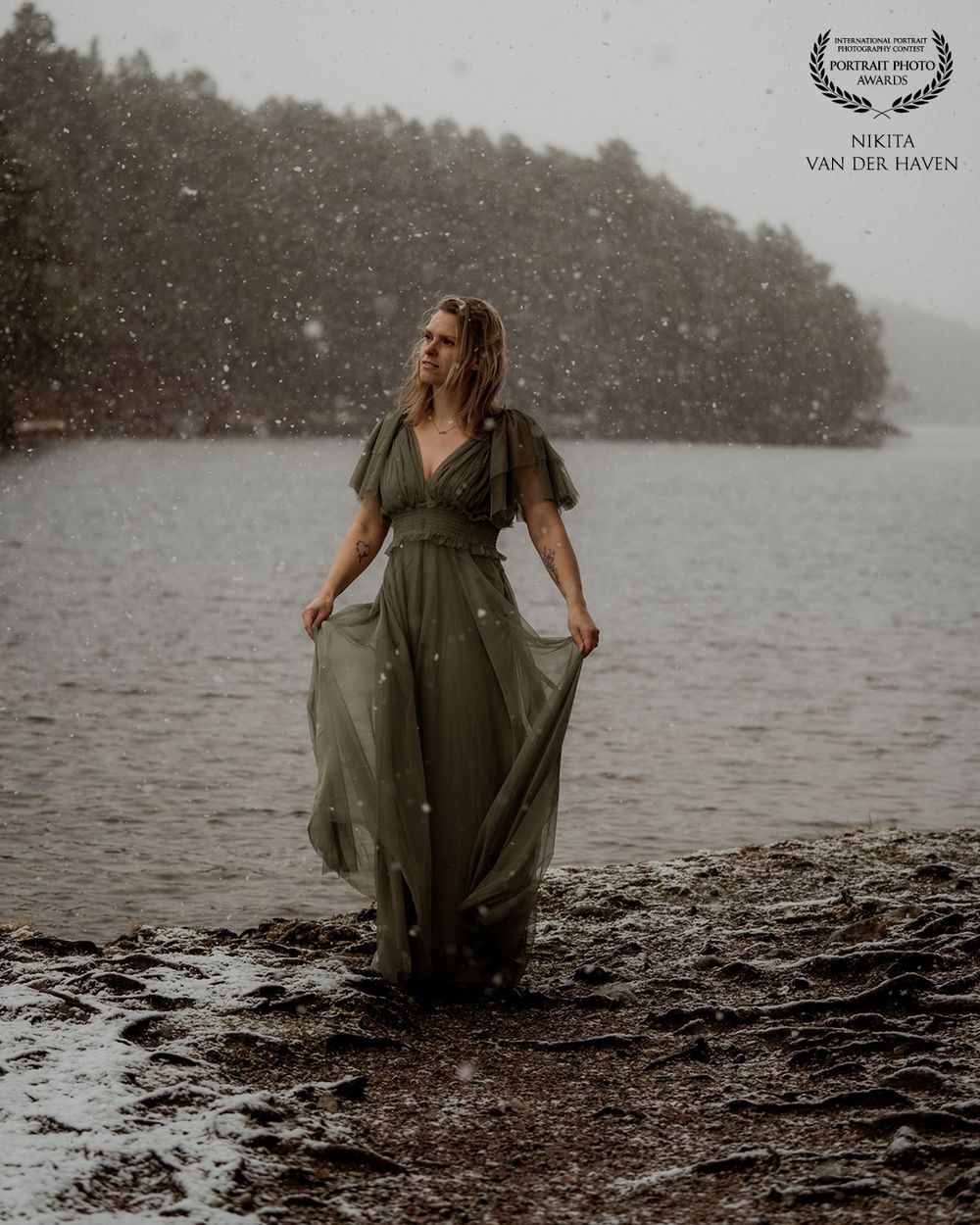 Woman in green dress standing in shallow water during snowfall with misty forest background.