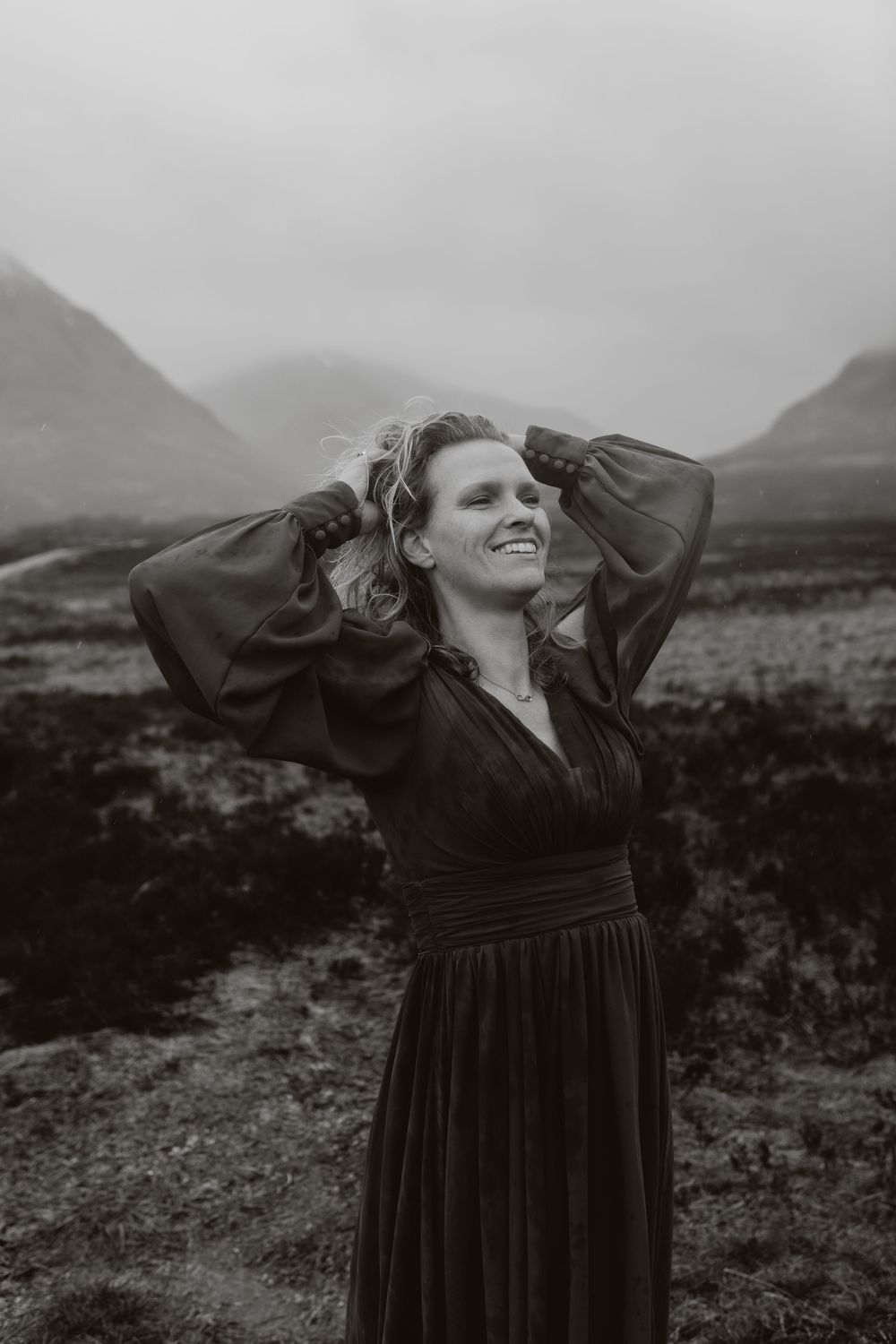 Woman in dark dress smiling with hands in hair, standing in misty mountain landscape, black and white photo.