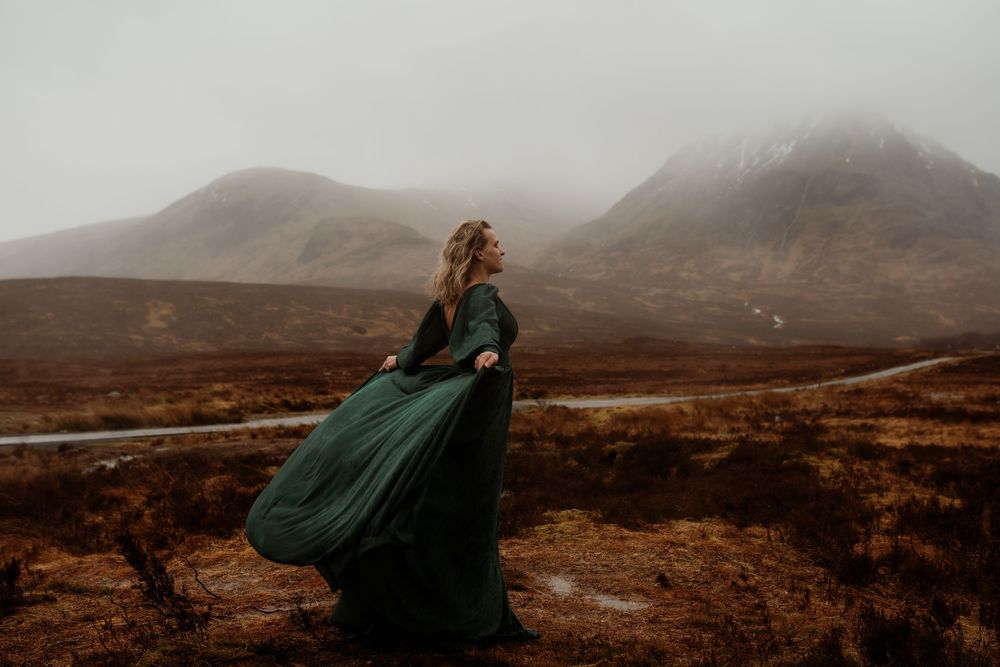 Woman in flowing green gown poses dramatically against misty Scottish Highland mountains.