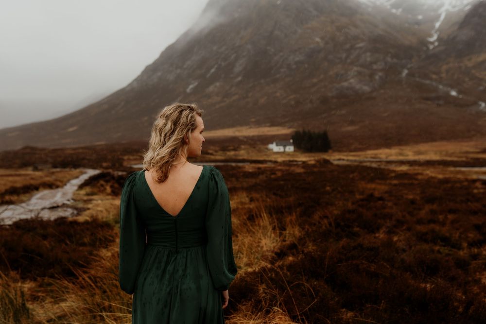 Woman in green dress gazes at misty Scottish highlands landscape with white cottage.