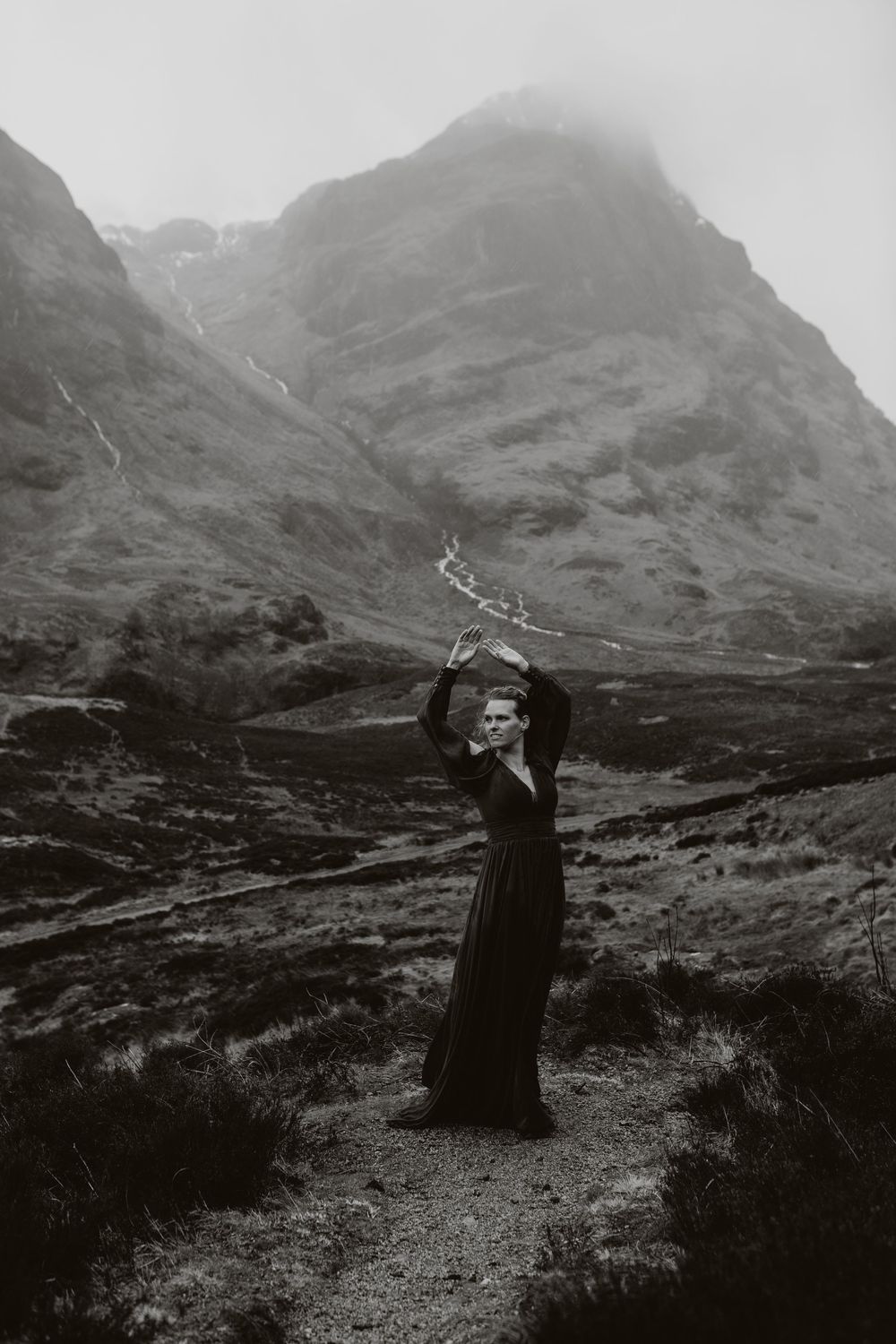 Black and white photo of woman in dark dress holding branches aloft in misty mountain valley.