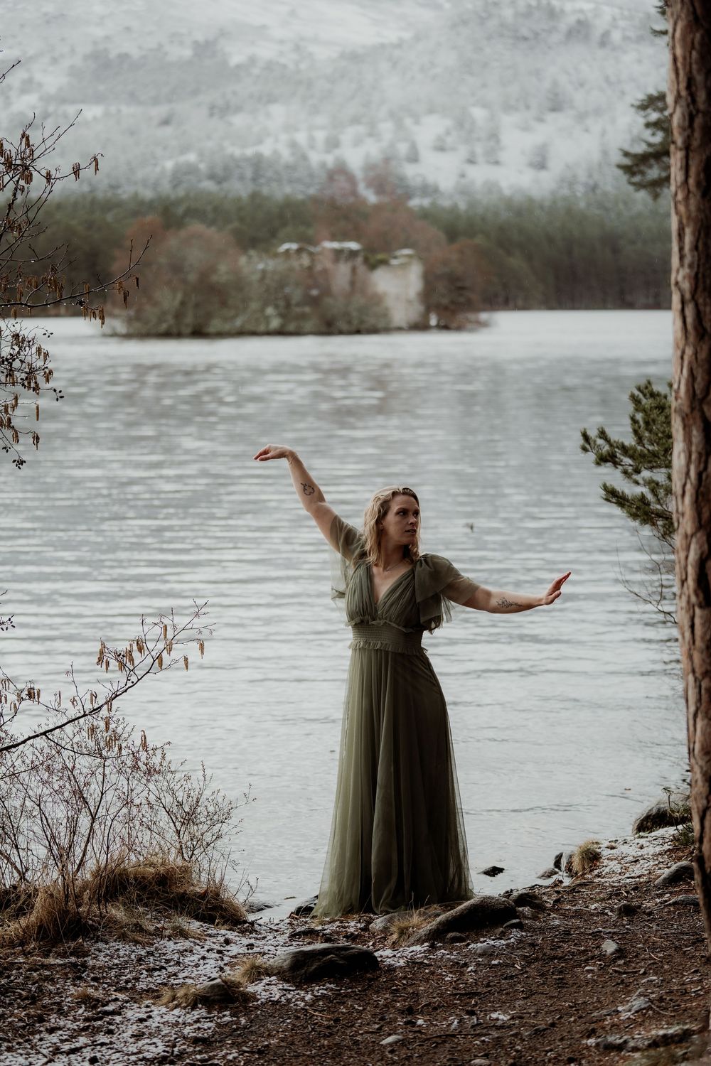 Woman in elegant olive green gown posing gracefully by a misty winter loch with snowy hills.