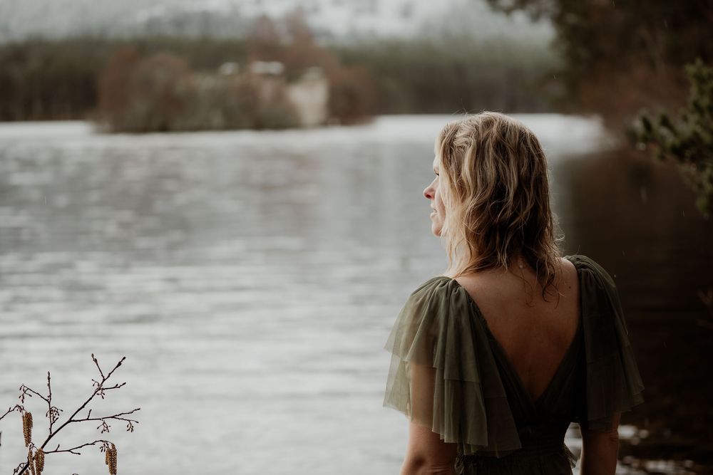 Woman in olive green backless dress gazing over a misty lake surrounded by bare winter trees.
