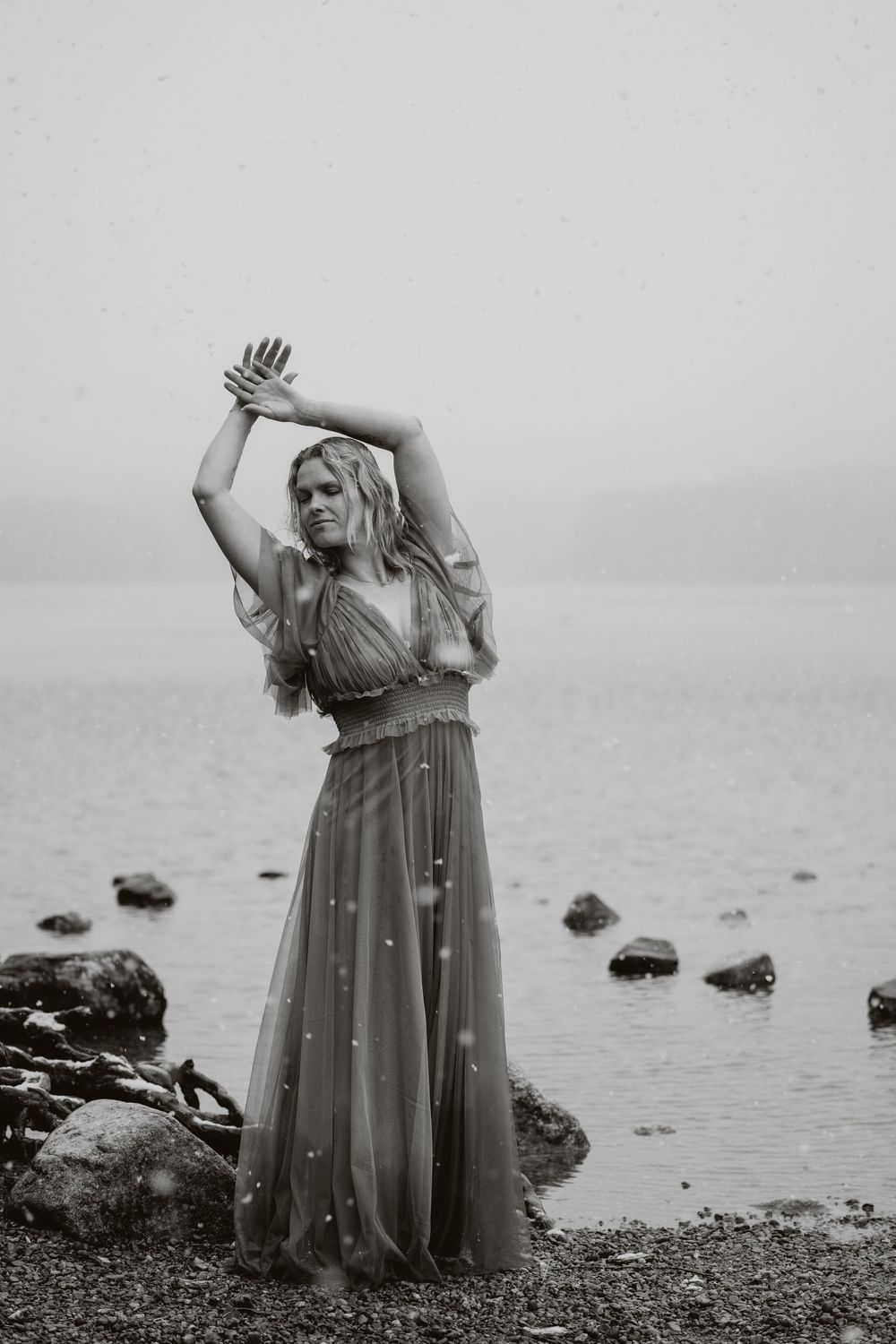 Woman in flowing gown poses gracefully on rocky shoreline in moody black and white photography.