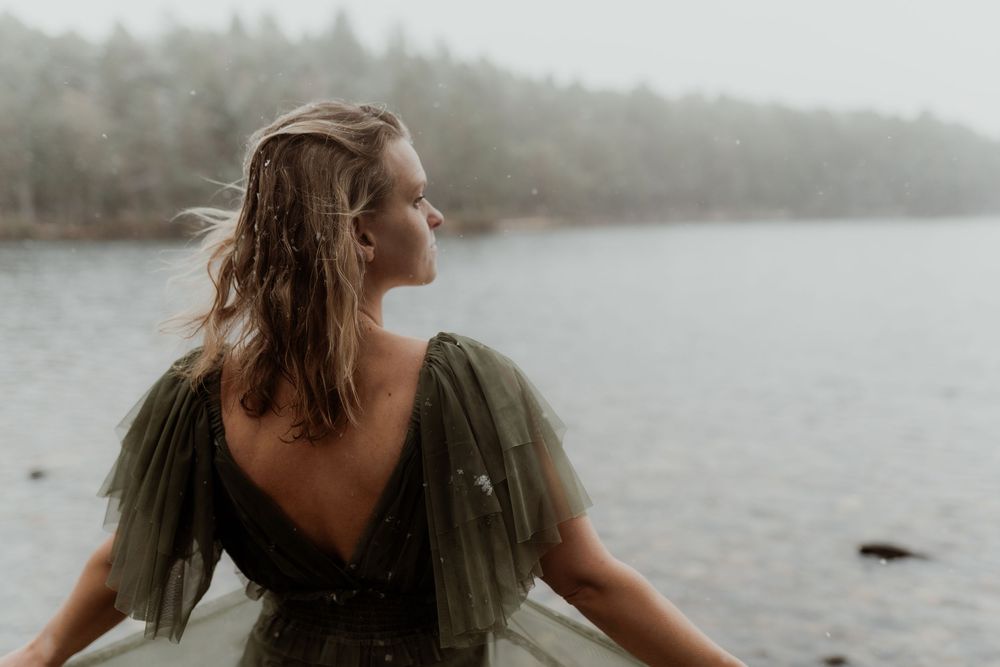 Woman in olive tulle dress stands by misty lake with wind-blown hair.