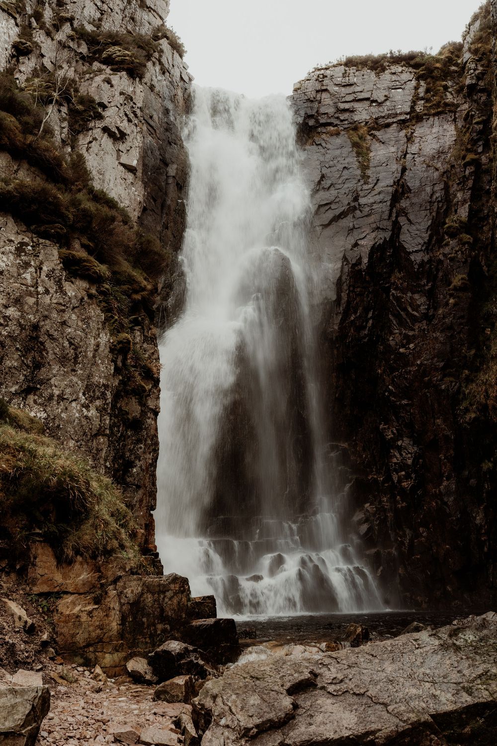 A powerful waterfall cascades down rugged rocky cliffs surrounded by moss-covered stone walls in a misty mountain gorge.