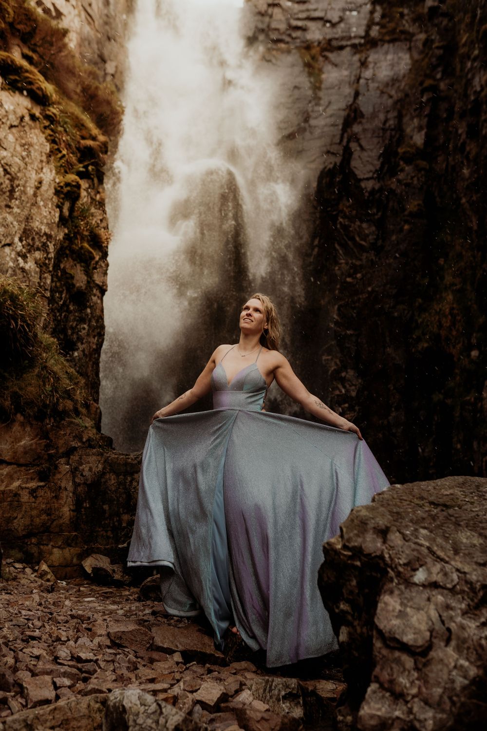 Woman in flowing iridescent gown poses dramatically before misty waterfall between rocky canyon walls.