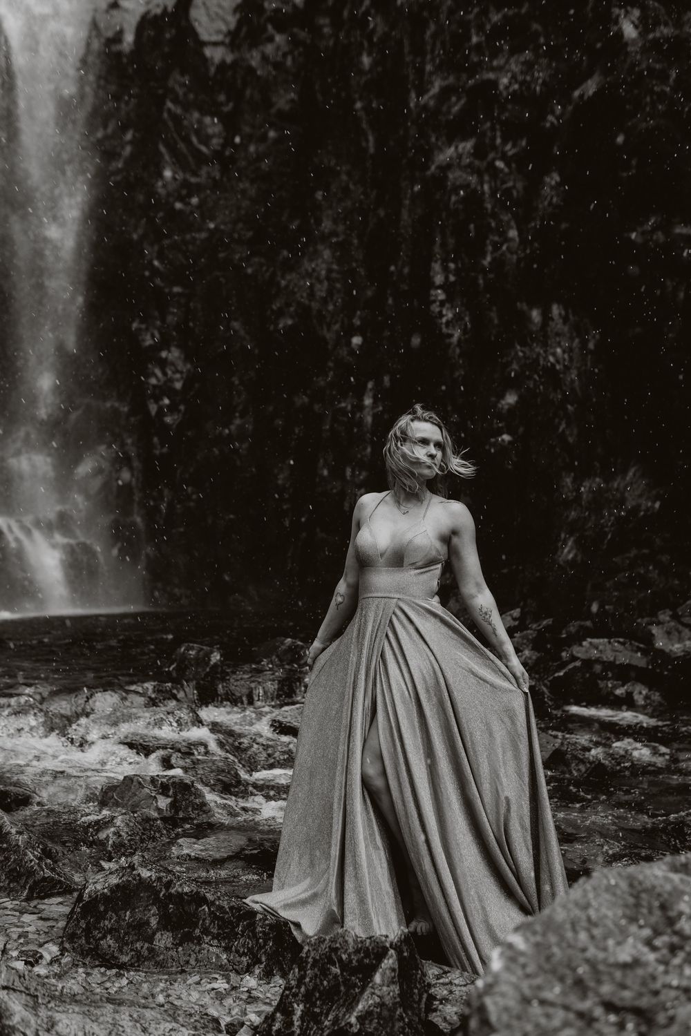 Black and white photo of a woman in an elegant gown standing before a waterfall on rocky terrain.