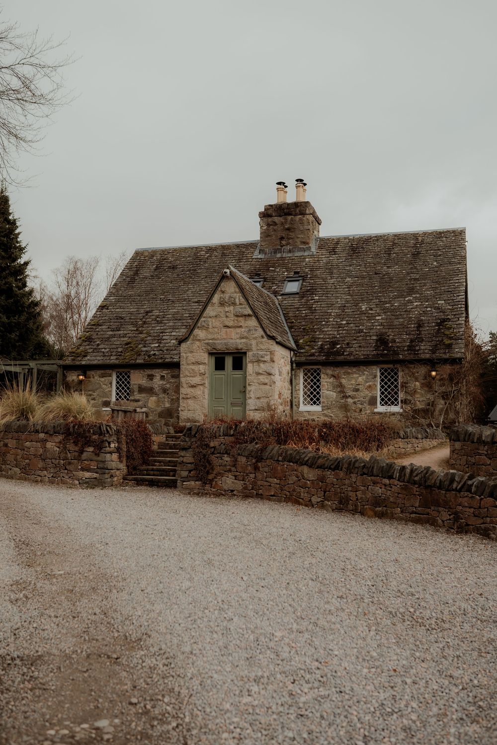 Charming stone cottage with green door and slate roof on a gravel path under overcast skies.