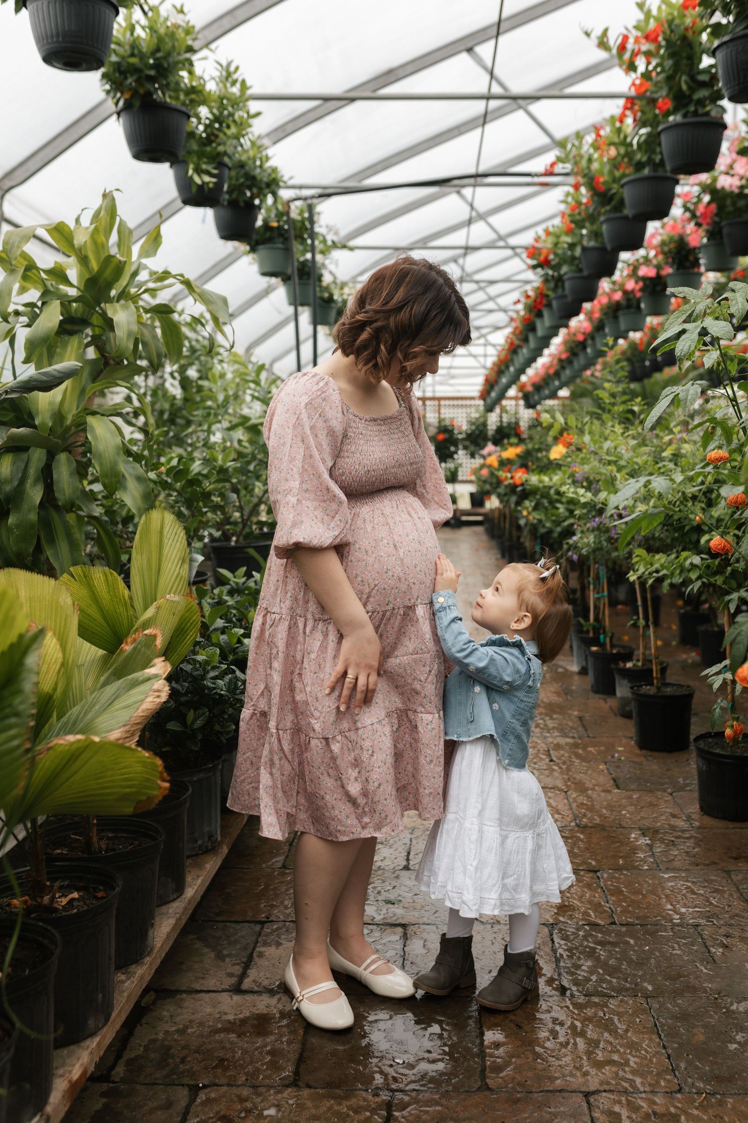 Pregnant woman in pink dress with toddler daughter in greenhouse surrounded by plants.