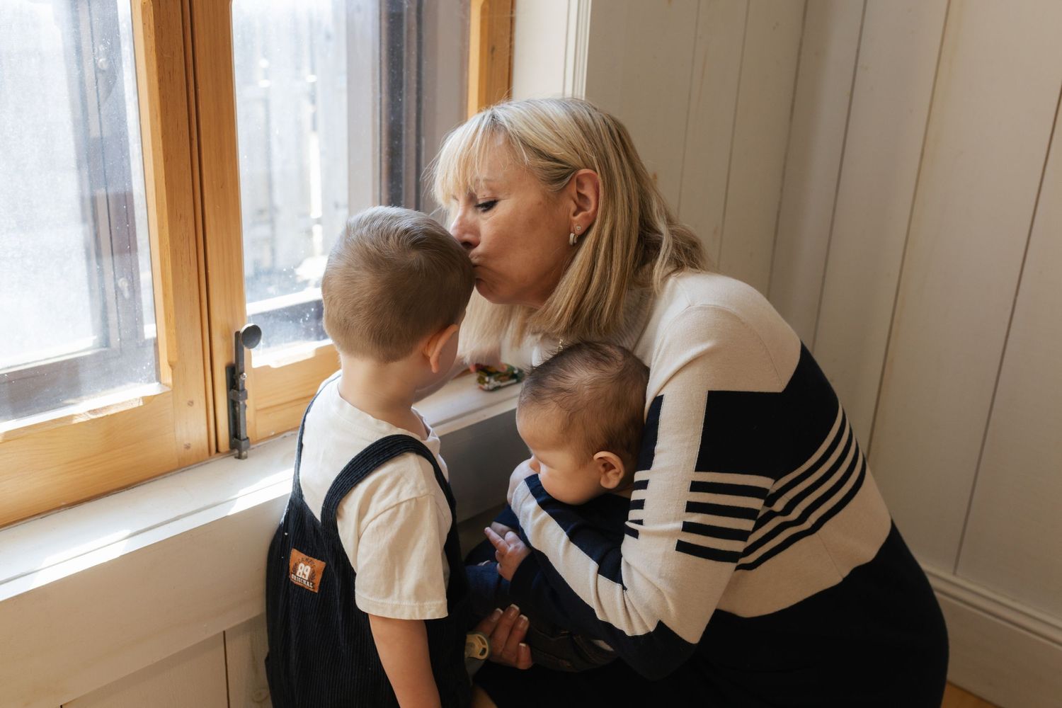 Loving blonde mother kissing young son while holding baby near window in cozy home interior.