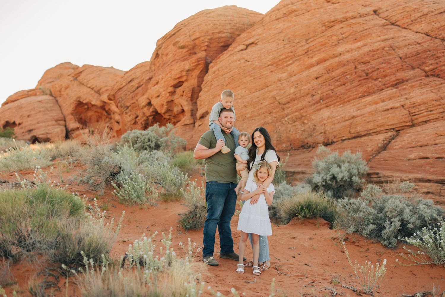 Family posing in front of red rock formations in a desert landscape with sparse green shrubs.