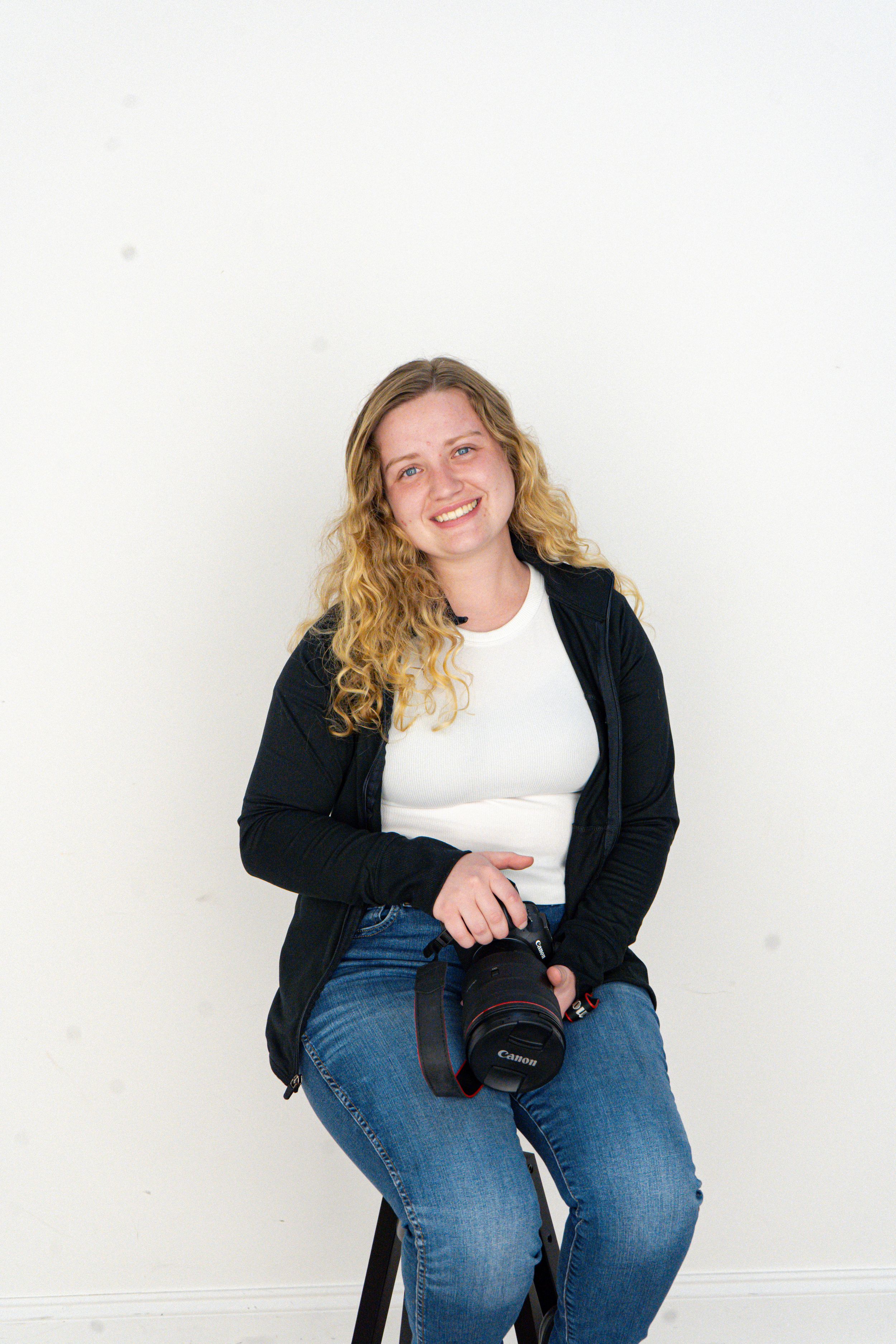 Smiling blonde woman holding a Canon camera, wearing jeans and black cardigan, posed against a white wall.