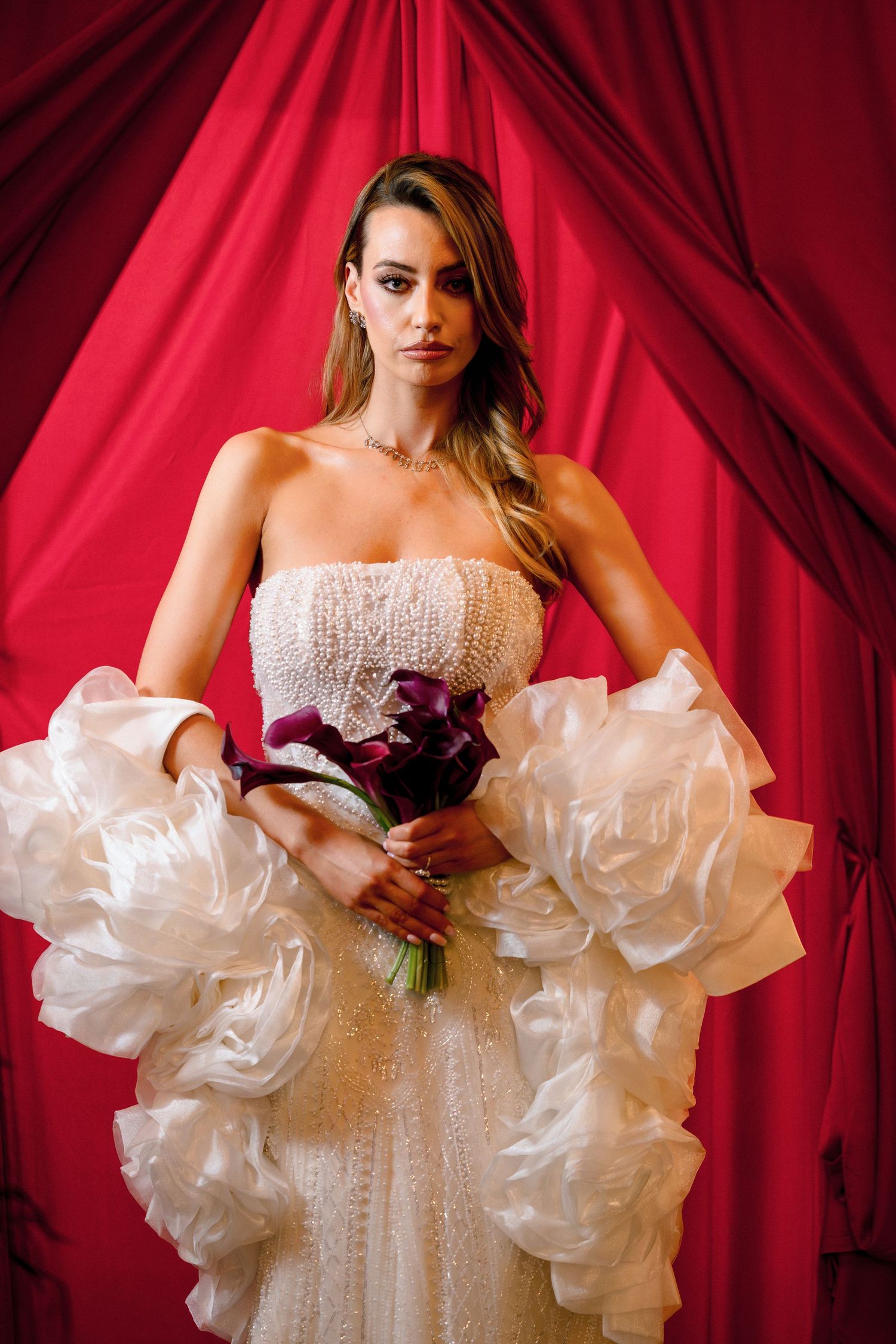 Bride in strapless beaded gown with white ruffled sleeves holds dark bouquet against red curtain backdrop.
