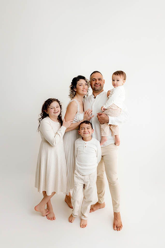 Family of five posing together in matching white and cream outfits against a white studio backdrop in Menifee CA.