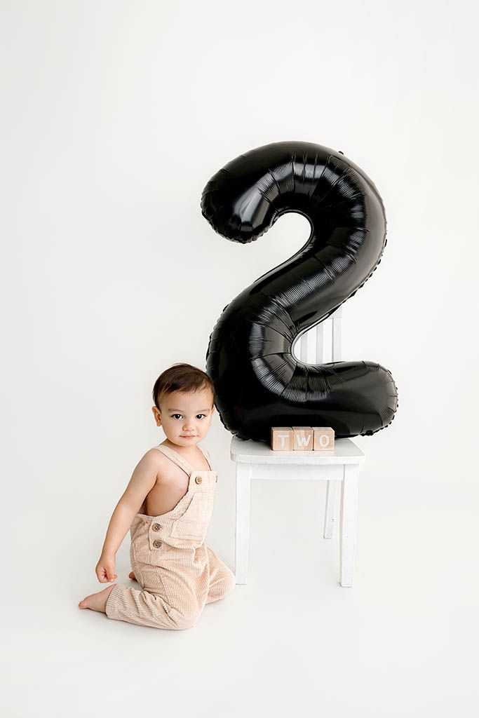 Toddler in beige overalls sits beside a white chair with a black number 2 balloon for a second birthday photo.