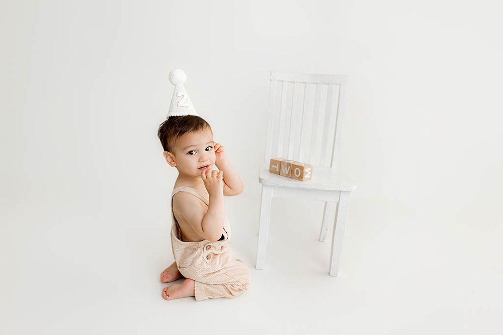 2nd birthday photographer in Menifee: Cute little boy in birthday hat sits on white floor near wooden blocks.