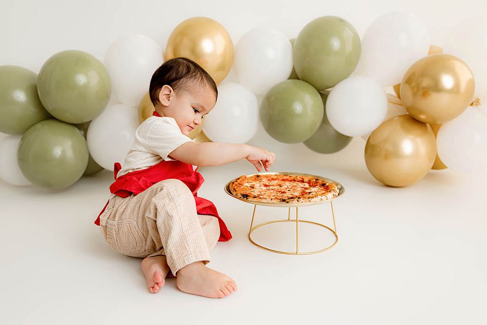 Toddler boy in a red apron reaches for a pizza on a gold stand surrounded by green and gold balloons.