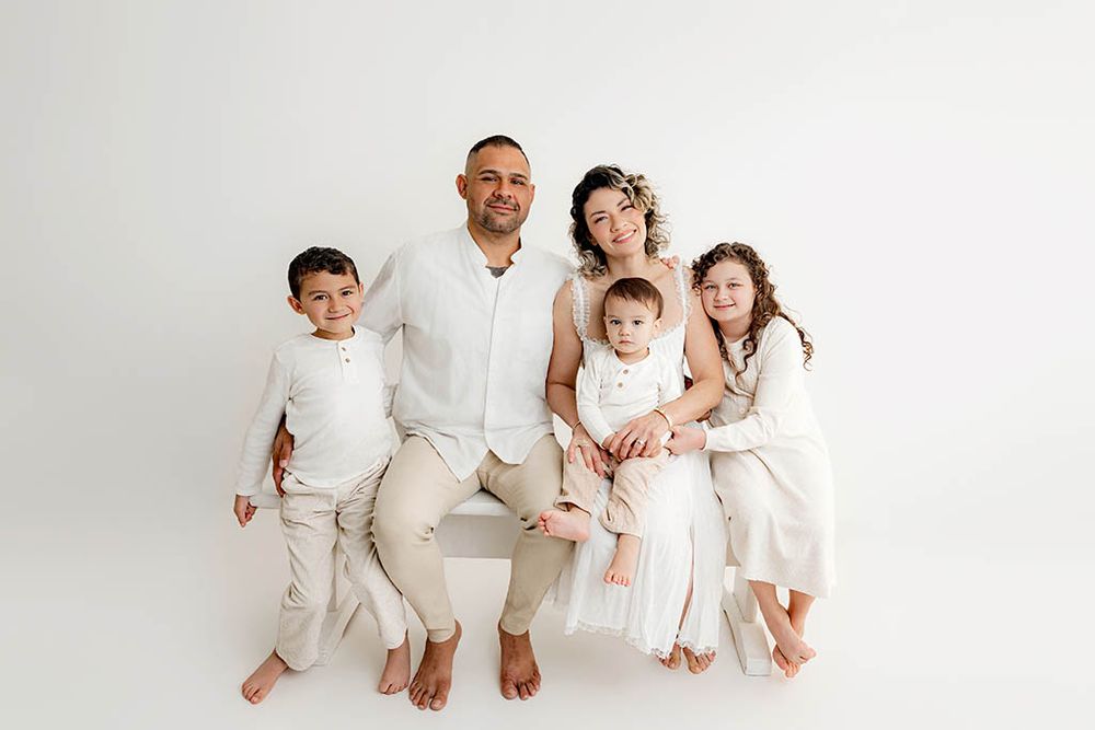 Family of five in matching white outfits posing together on a bench against a white studio background.