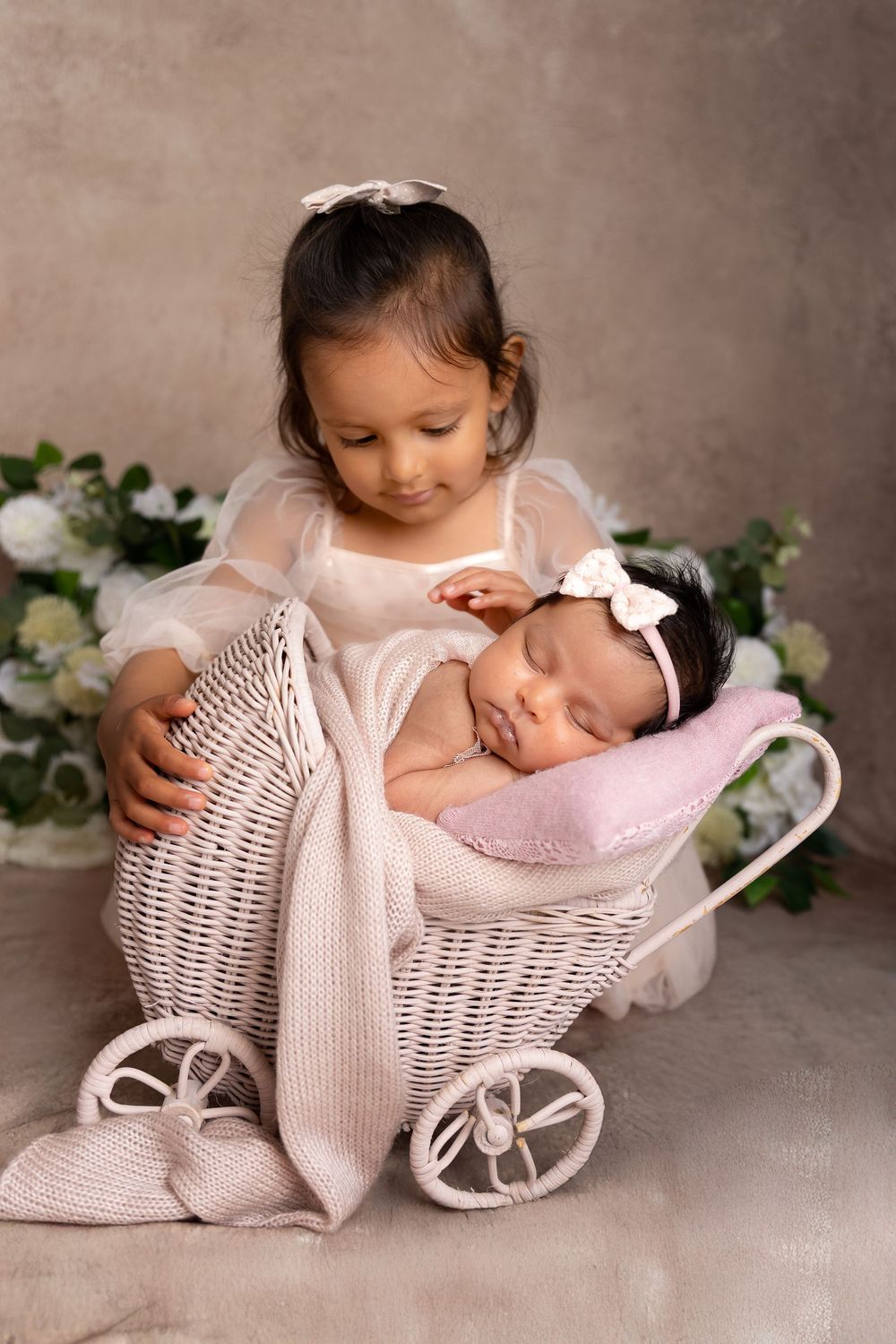 A toddler girl in white dress lovingly holds a sleeping newborn baby in a white wicker pram during a studio photo session.