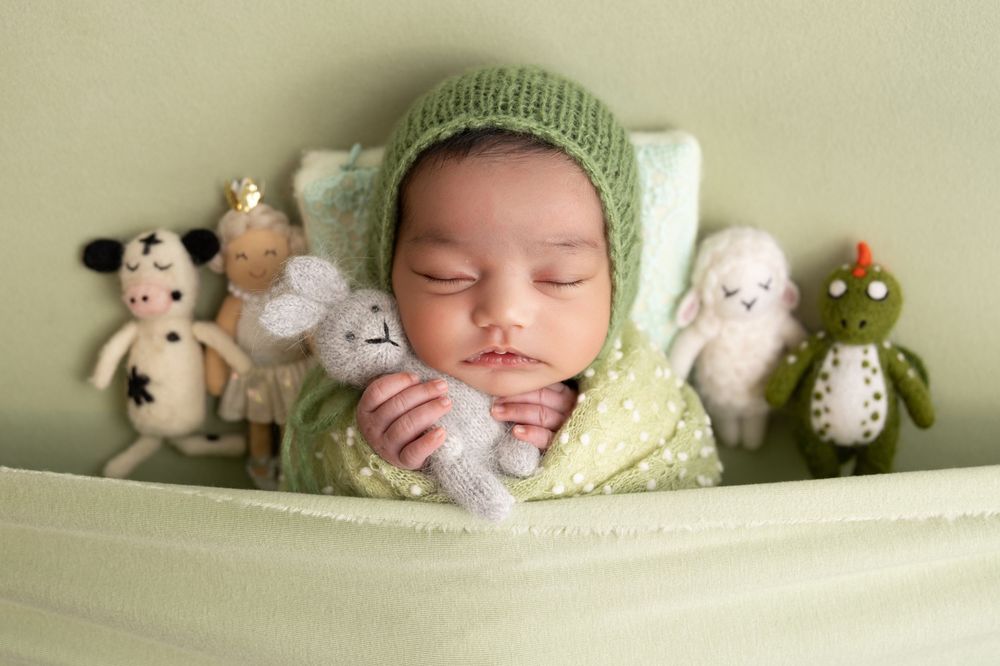 Sleeping newborn baby in green knit bonnet surrounded by stuffed animal toys on a soft green background.