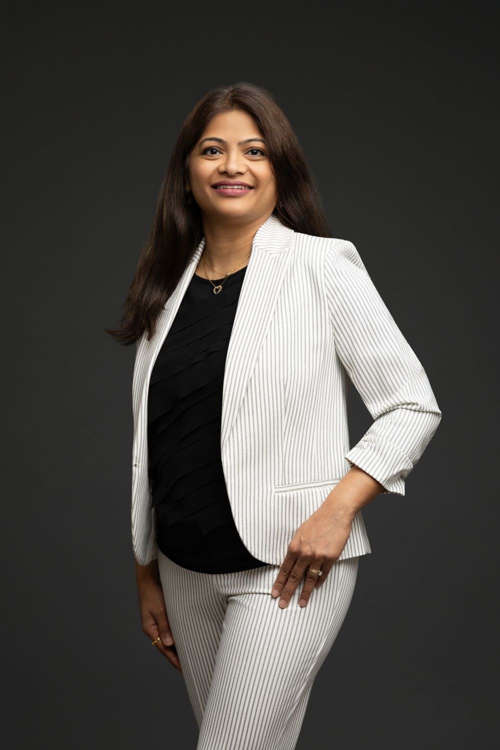 Professional woman in white pinstripe blazer and pants poses confidently against dark gray studio background.