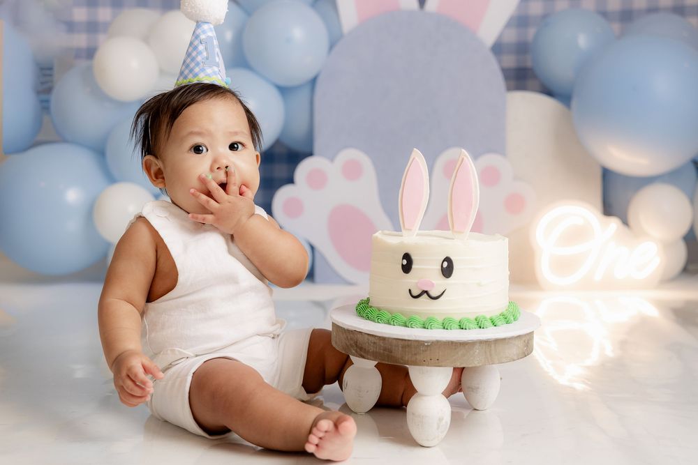 Baby in white outfit enjoys first birthday cake smash with bunny cake and blue balloon backdrop.