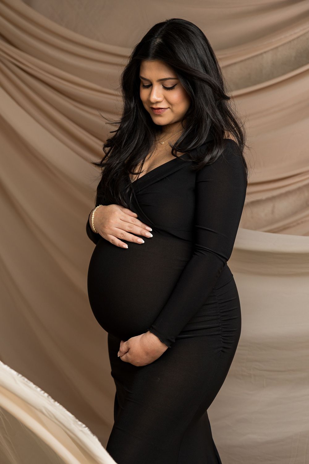 Pregnant woman in black dress cradling baby bump against beige draped backdrop in maternity photo.