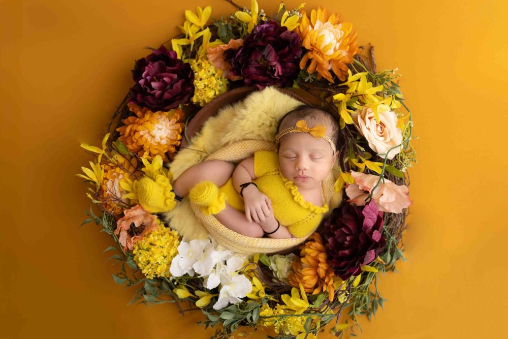 Newborn baby sleeping in a wooden bowl surrounded by a colorful floral wreath on a golden yellow background.