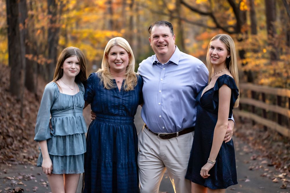 Family of four smiling outdoors on a wooden bridge surrounded by colorful autumn foliage.
