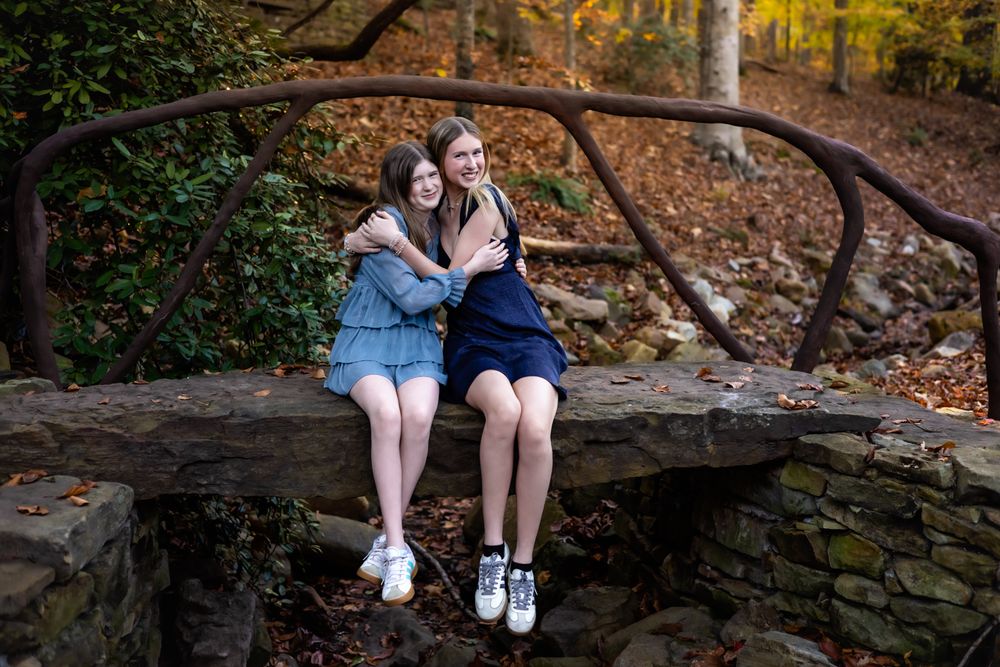 Two teenage girls hugging and smiling on a stone bridge surrounded by autumn foliage in a forest.