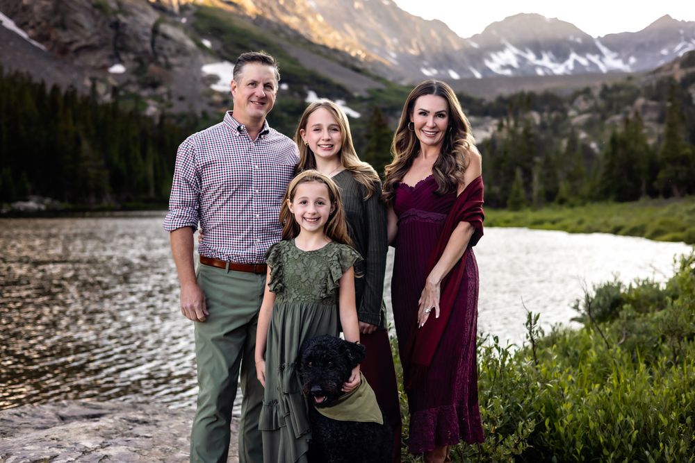 Family of five posing near a mountain lake with snow-capped peaks in the background during golden hour.