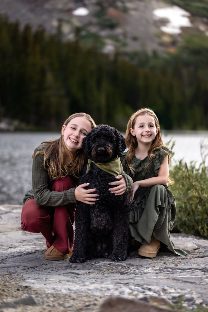 Two smiling girls pose with a black curly dog by a mountain lake, surrounded by pine trees.