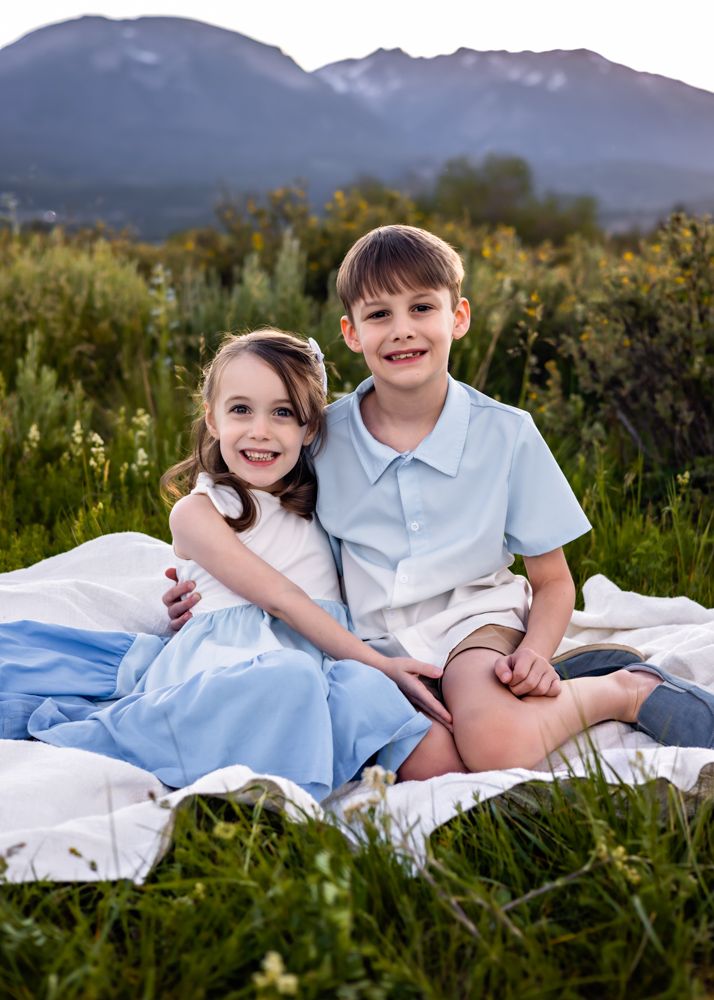 Two smiling children in blue and white outfits sit on a blanket in a mountain meadow during golden hour.