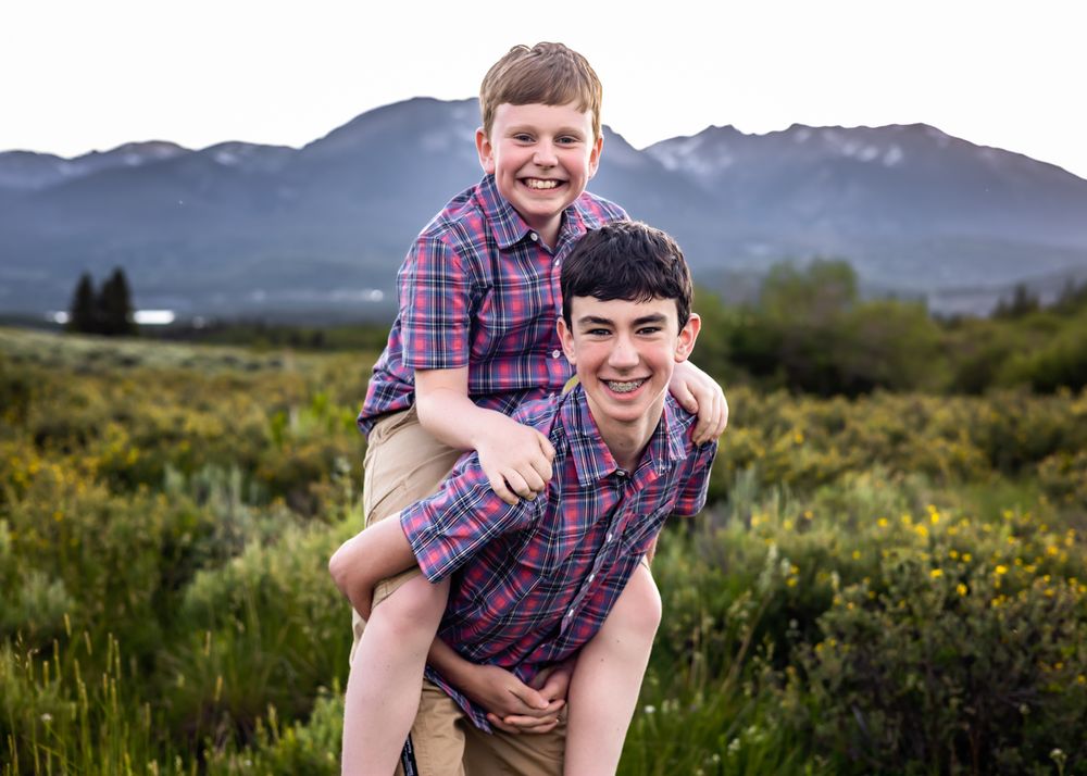 Two smiling boys in matching plaid shirts pose piggyback in a mountain meadow with green shrubs and peaks behind them.