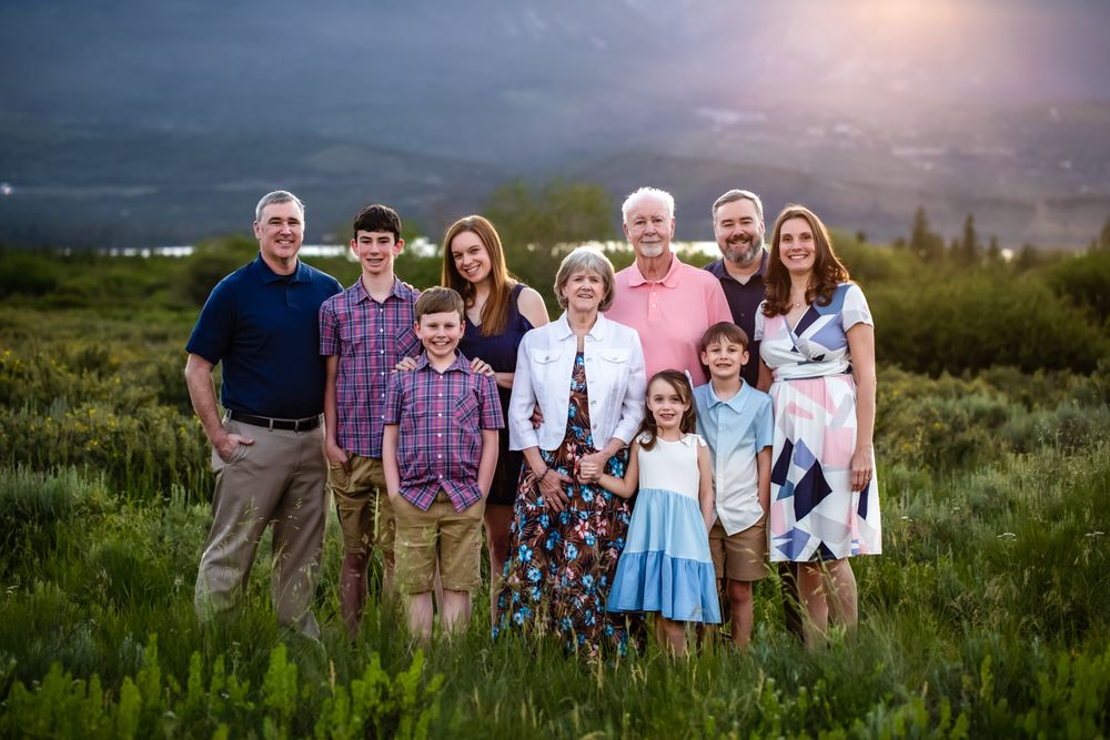 Multi-generational family portrait outdoors in lush green meadow with dramatic stormy sky backdrop.