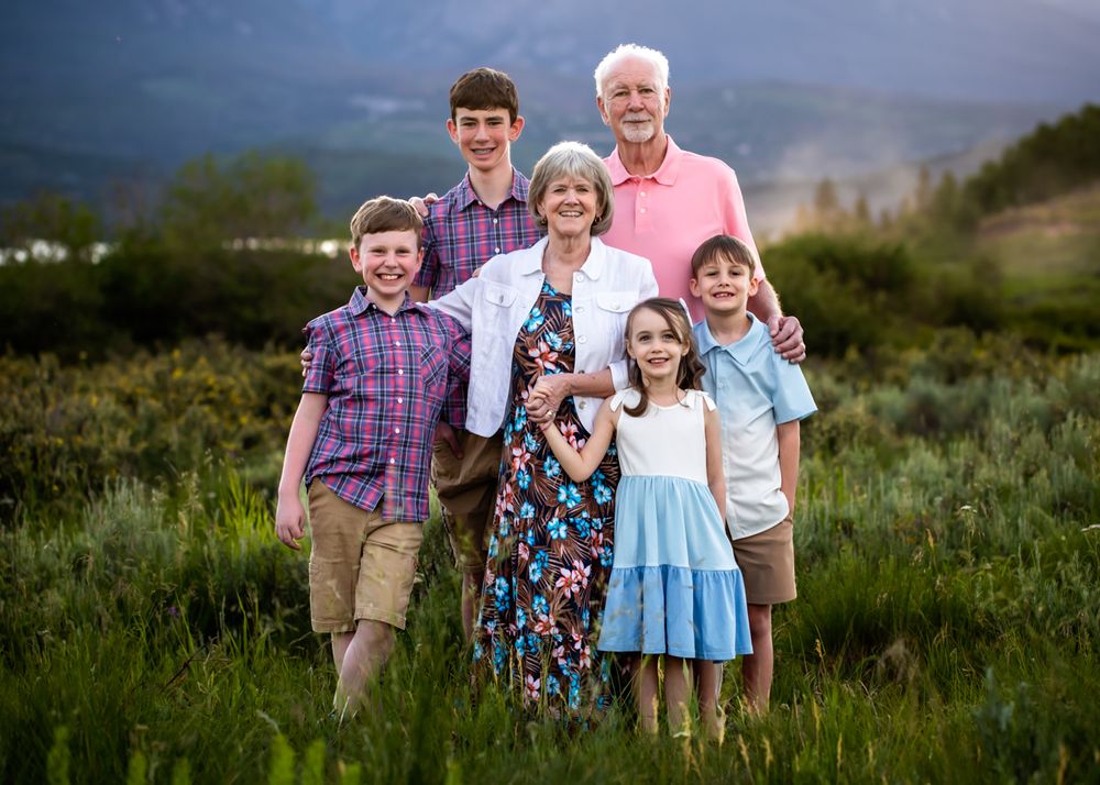 Family of six posing outdoors in a lush green field with mountain backdrop during golden hour.