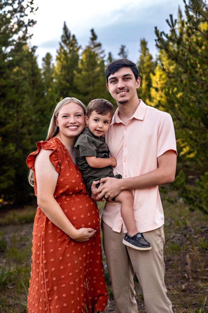 Smiling pregnant woman in red dress with husband holding toddler son, posing outdoors among green pine trees.