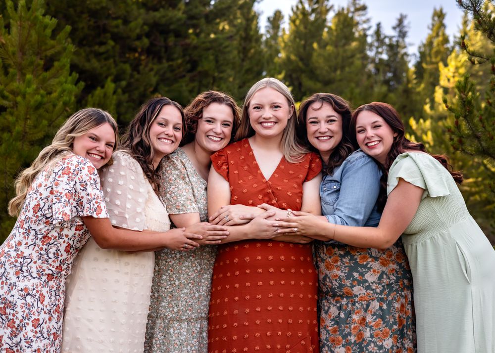 Six smiling women in floral dresses pose together outdoors with pine trees in the background.