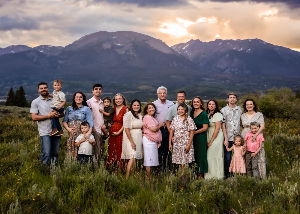 Large family portrait outdoors with mountain sunset backdrop, adults and children smiling in coordinated outfits in a meadow.