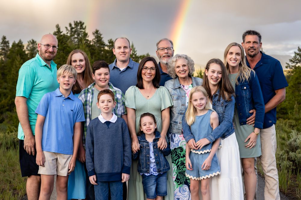 Large family group portrait smiling outdoors with a vibrant rainbow arching over pine trees in the background.