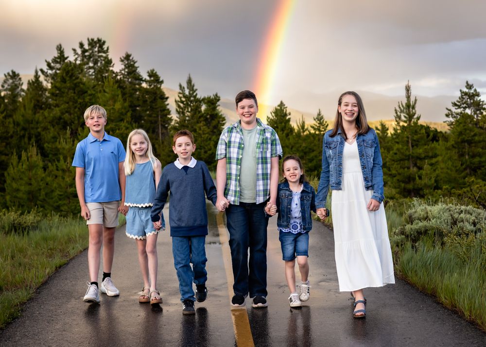 Six children walk hand-in-hand on a road with a vibrant rainbow over lush green trees in the background.