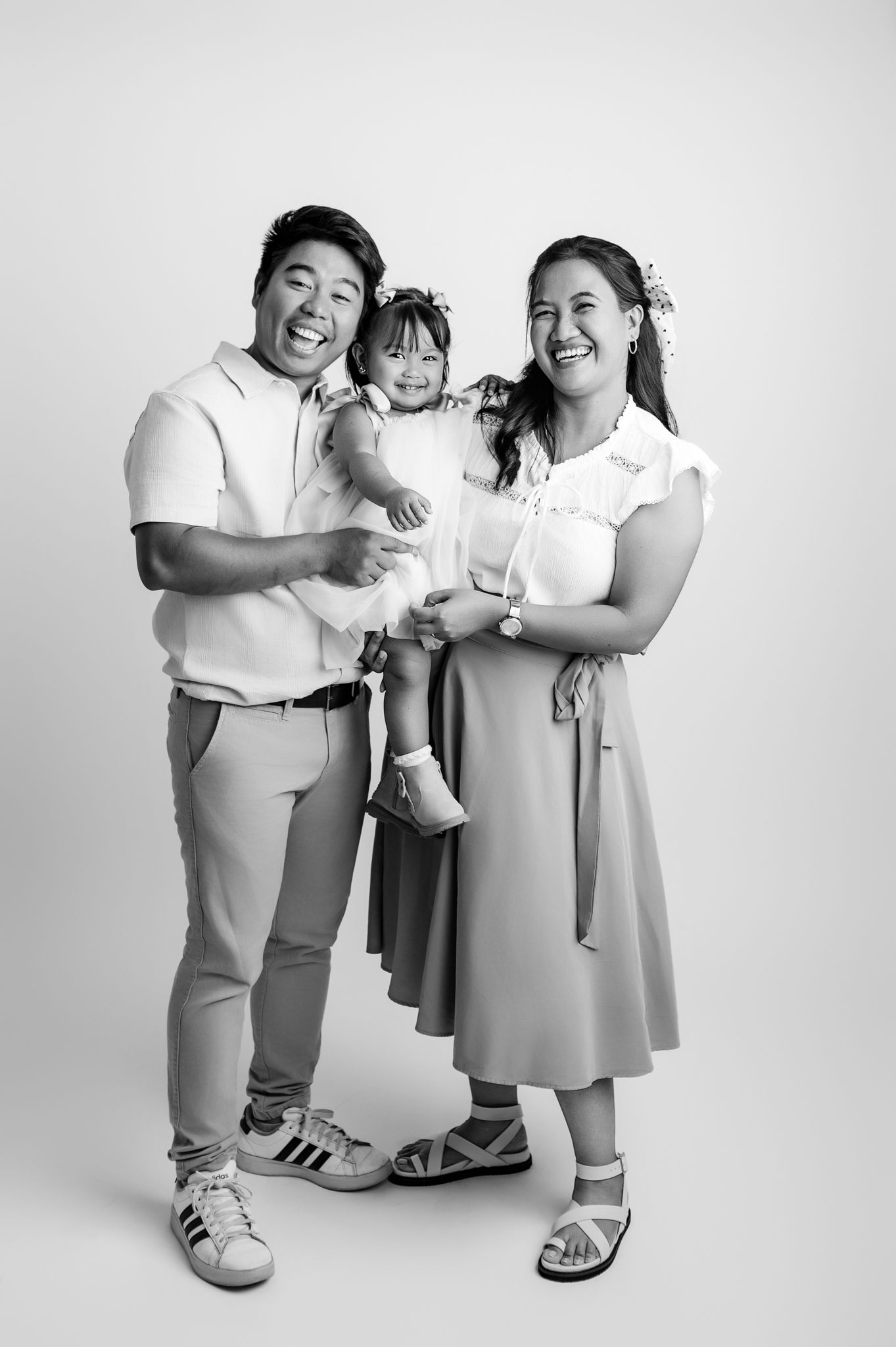 Black and white studio portrait of smiling Asian family with parents holding young toddler daughter in matching outfits.