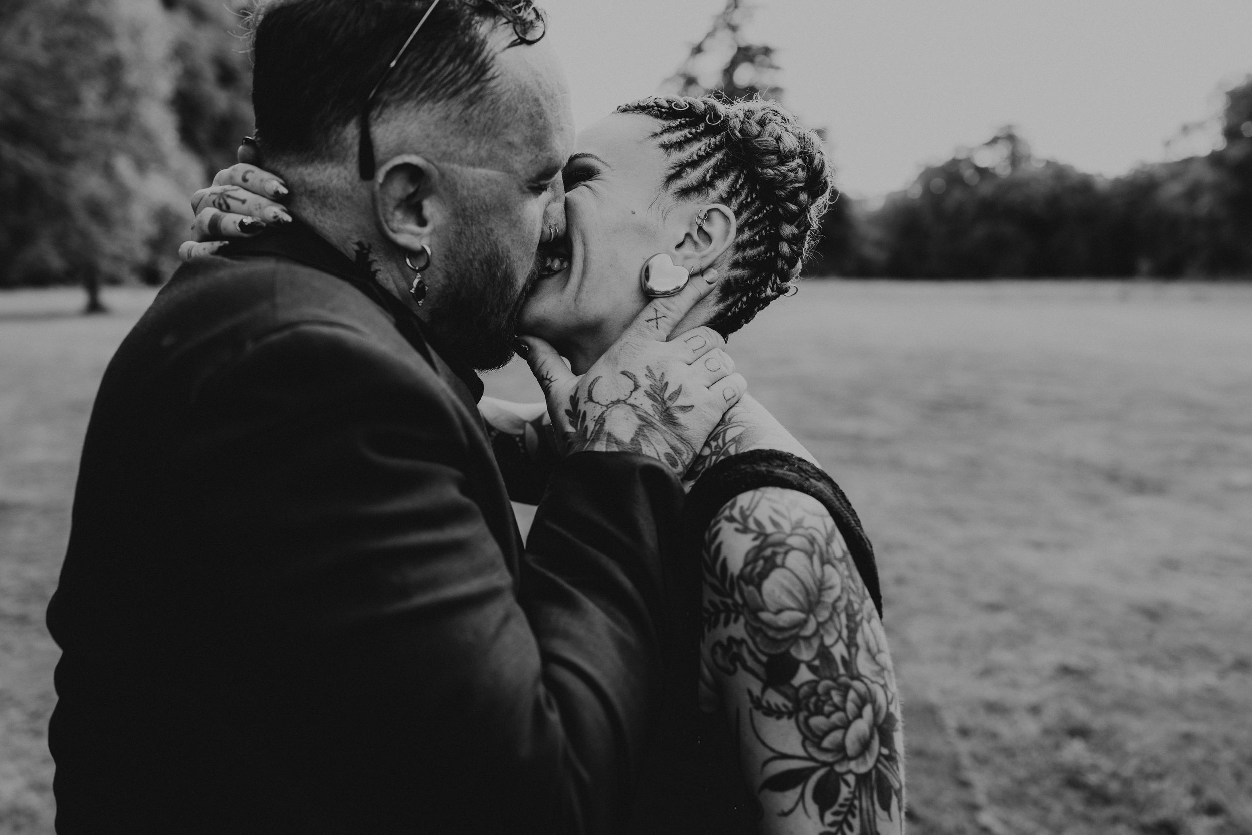 Black and white photo of tattooed couple sharing a romantic kiss outdoors in a park setting.