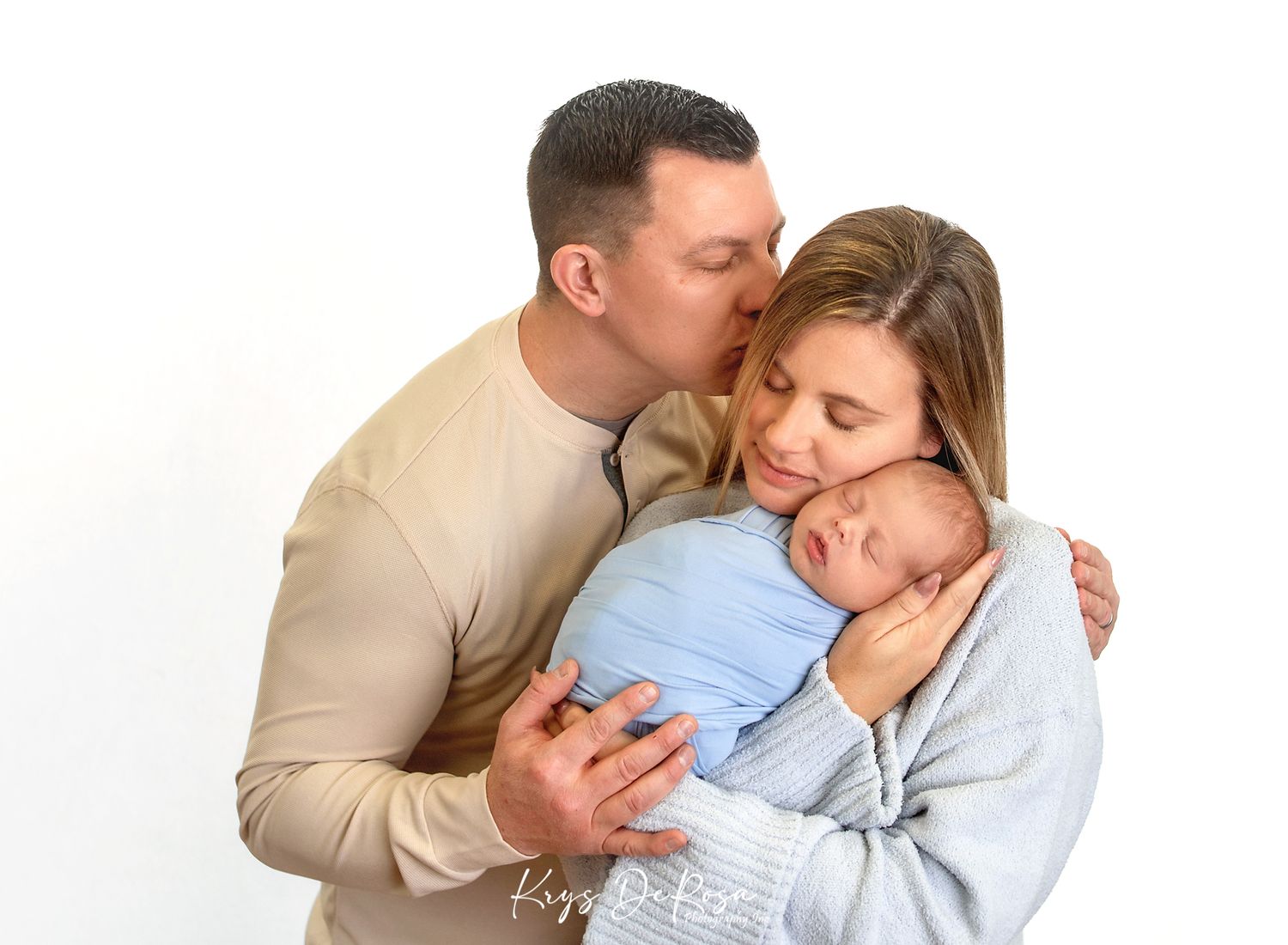 Parents sharing a tender moment, father kissing mother's cheek as she cradles their sleeping newborn baby wrapped in blue.
